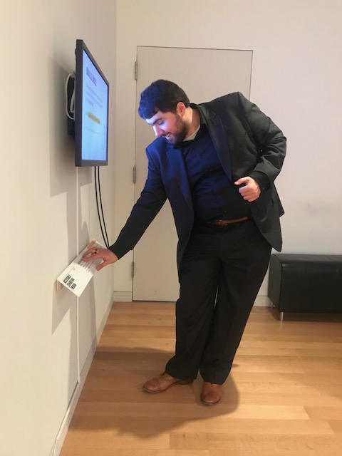 Sina stands in front of a video screen in a gallery bending over slightly to read the braille on a label mounted on an angle coming off the wall.