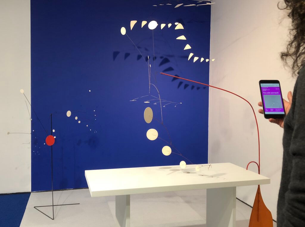 A grouping of Alexander Calder's mobiles and stabiles is depicted. On the right side of the image, a curly haired person, who is mostly cut off from the image, holds a phone up in front of the works.