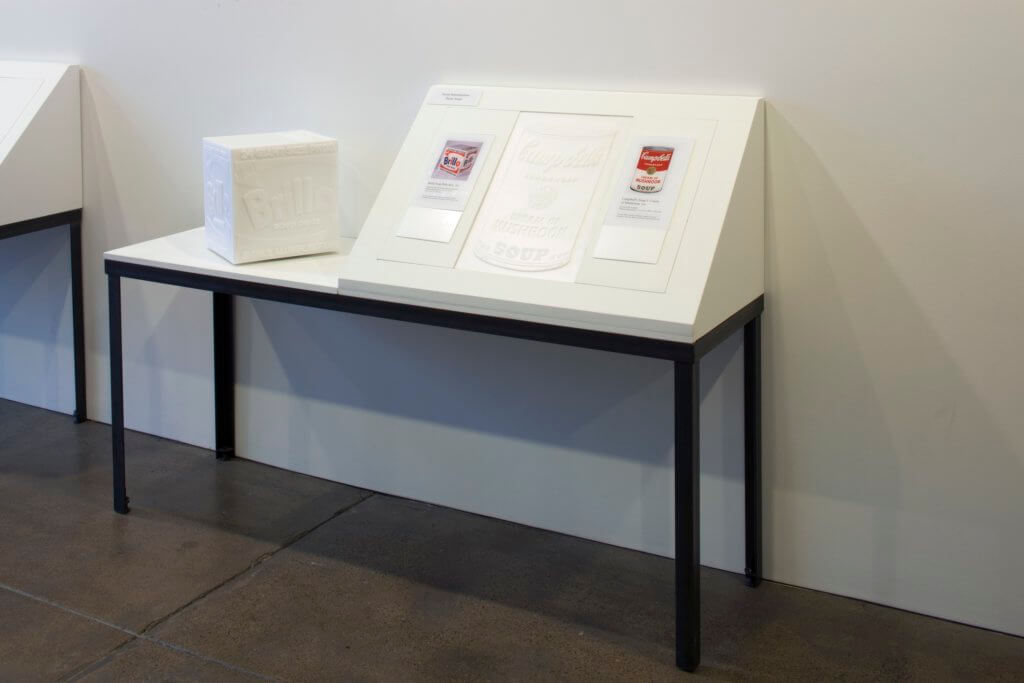 A tactile replica of a Brillo Soap Pads Box sits on a table next to an angled display featuring a tactile reproduction of a Campbell's Soup can and small prints of the artworks.