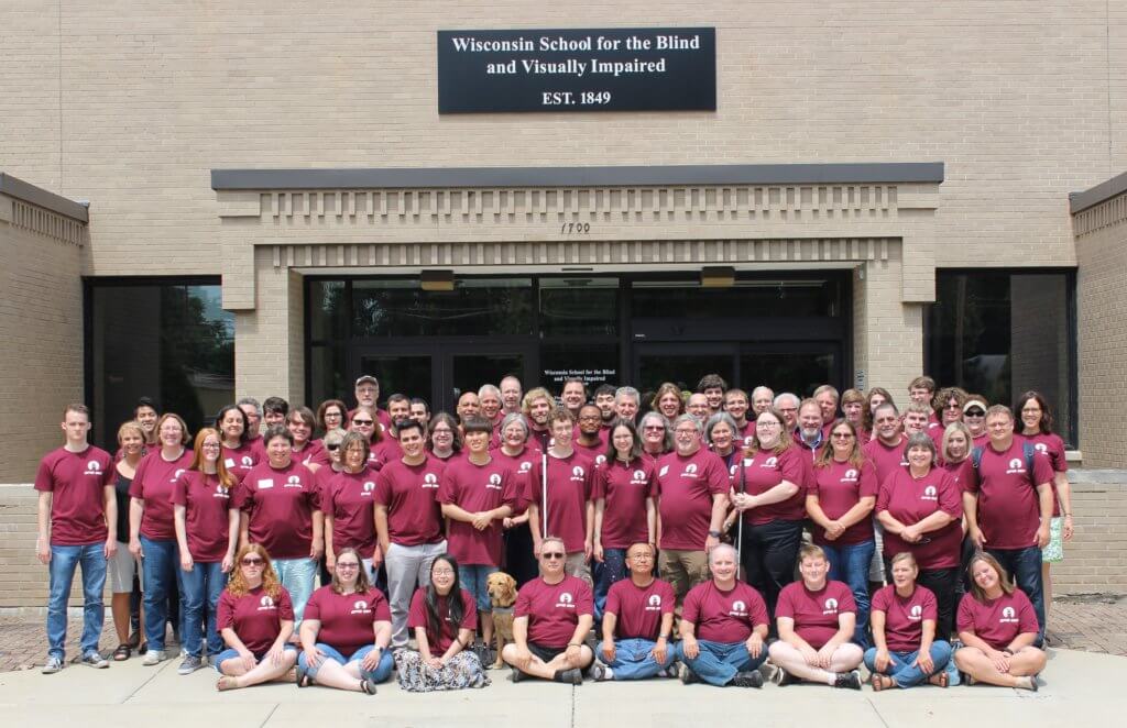 A group of 70 people in maroon shirts pose in front of the entrance of WSBVI.