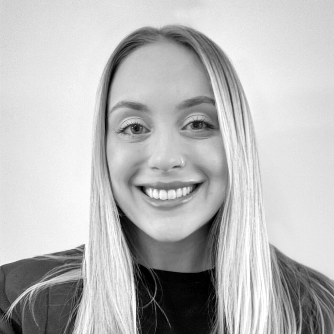 A black and white headshot of a young white woman with light skin and a wide toothy smile. Shown from the chest up, she has long straight blonde hair, parted in the middle that frames her face. She looks directly at us with light eyes, wears a small silver nose ring, and has defined high arching eyebrows. She wears a black top under a dark grey blazer in front of a blank wall.
