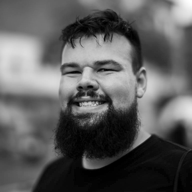 A black and white close-up outdoor headshot of a White adult man with a big, wide smile looking directly at us. He has dark hair styled slightly tousled with shorter sides, dark eyes, thick eyebrows, and a full beard and mustache. His face is broad with full cheeks that stand out while smiling. He is wearing a dark T-shirt. The background is softly blurred, with indistinct buildings and foliage.
