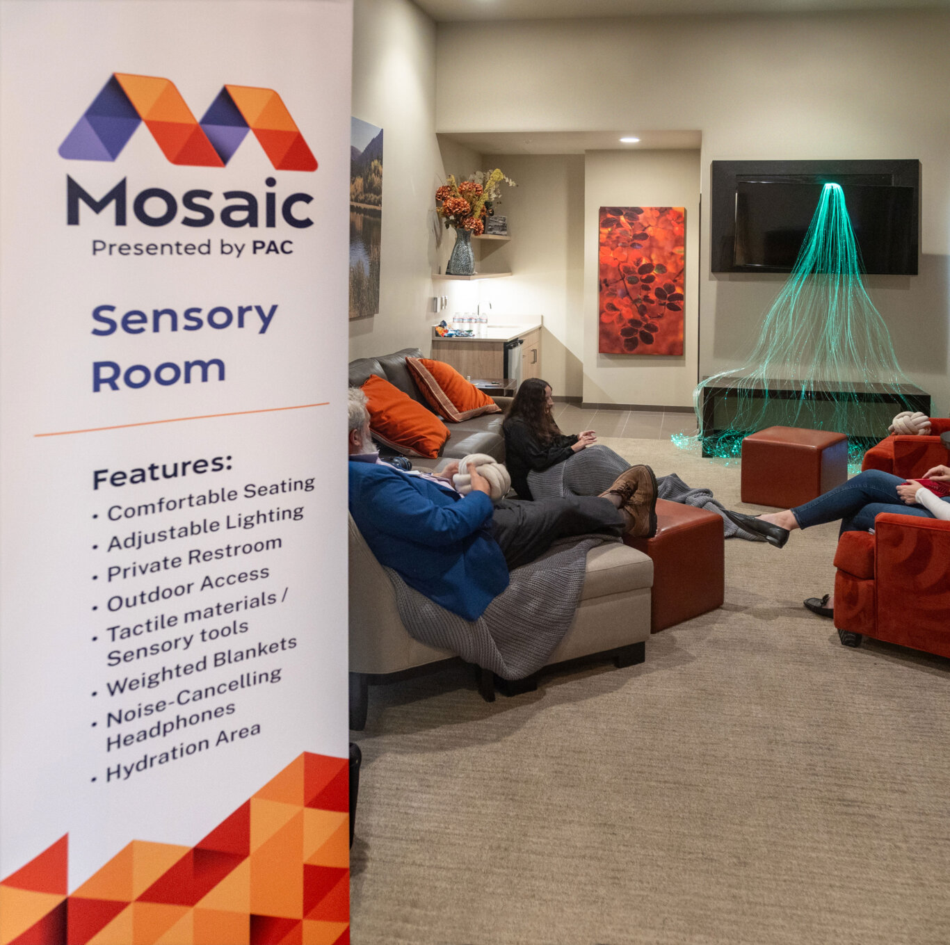 The entrance to a sensory room. On our left, a large vertical sign reads "Mosaic Presented by PAC" with the Mosaic logo and sub heading "Sensory Room." Underneath that is a list of features, "comfortable seating, adjustable lighting, private restroom, outdoor access, tactile materials/sensory tools, weighted blankets, noise-cancelling headphones, and a hydration area." In the background, people with light skin tone are seated on couches and the floor with orange pillows and blankets playing with various fidgets. The room includes wall art, a small kitchenette area, and a glowing green fiber-optic light feature cascading over the wall mounted TV.