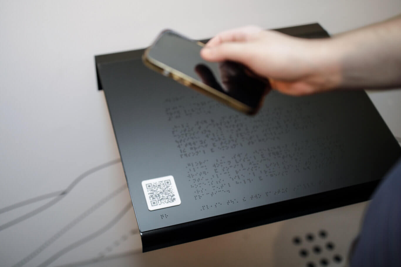 A shallow-focus, slightly blurry close-up of a hand with light skin holding a smartphone above a white tactile QR code that sits in the lower left hand corner of a black reader raille covered in Braille. Beneath the reader rail on the ground, out of focus are a series of raised tactile floor markers.