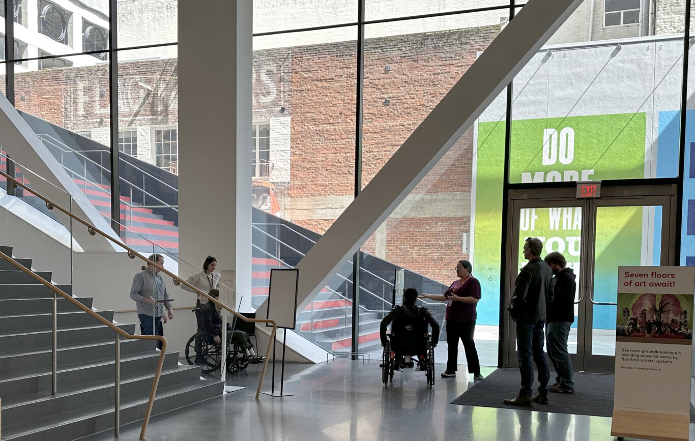 A spacious, modern art museum lobby filled with natural light from tall floor-to-ceiling windows. Several people, silhoetted against the windows, are gathered near the entrance: some are standing and talking, and two people are using wheelchairs. Large white structural beams and a glass-railed staircase dominate the interior, while another staircase with red accents is visible outside through the windows. Beyond the glass is a brick building wall. On our right, there are glass doors with an illuminated red "EXIT" sign above them, a colorful green-and-blue wall graphic with partial text, and a sign reading "Seven floors of art await!" The polished floor reflects the light and figures.