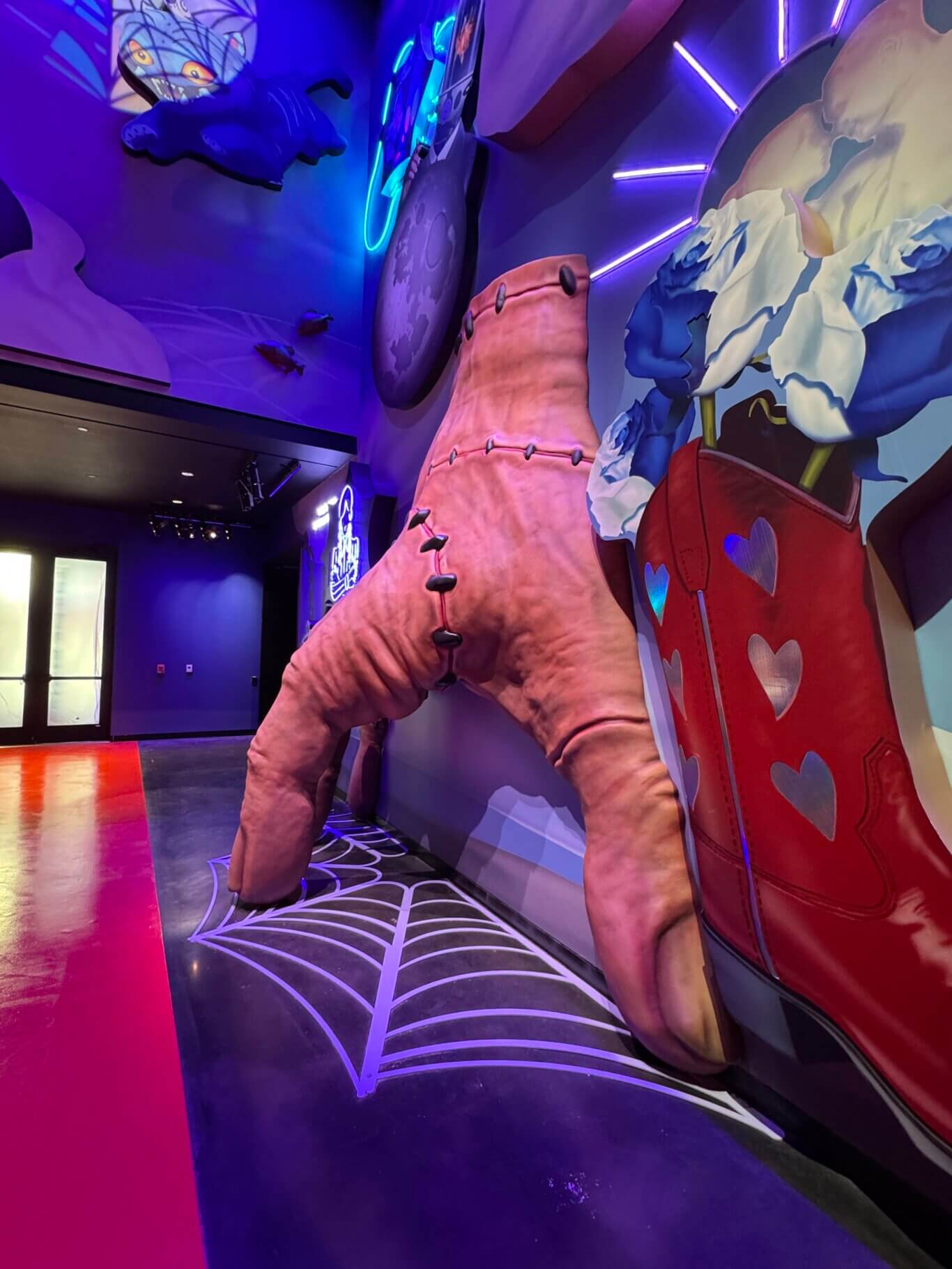 A colorful themed interior space dominated by vivid purple, blue, and pink lighting of a towering replica of a dismembered hand, fingertips on the ground as if skittering out of the wall on our right. The hand, a representation of Thing from Wednesday, has light skin tone and large stichings stretching across scars. The floor has a large white tactile spiderweb pattern. An oversized cartoon red cowboy boot with hearts is next to the hand.