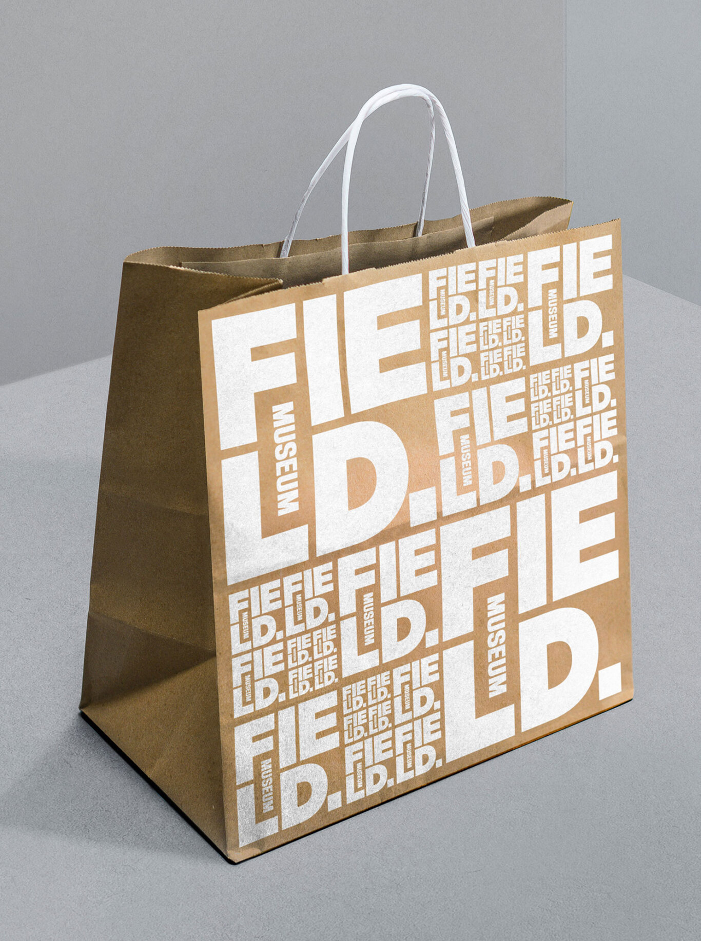 A brown paper shopping bag photographed in a studio-like setting. The bag is upright and slightly angled, revealing both the front and left side. Its front is covered with large white block typography repeating the words "FIELD." and "MUSEUM" in different sizes and orientations, creating a dense graphic pattern. Two white twisted paper handles arch above the open top of the bag. The background and tabletop are neutral gray, emphasizing the bag's brown paper texture and high-contrast white print.
