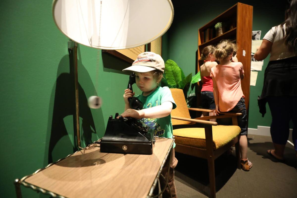 A young child with pale skin plays with an oldr rotary phone in a warmly lit, museum-like room with dark green walls and vintage furnishings. The child, wearing a cap and green shirt, holds the receiver of an old black rotary telephone while pressing or touching the phone's base on a wooden table. A large floor lamp with a white shade stands beside the table, casting a strong shadow on the wall. In the background, other children and an adult stand near a wooden shelving unit, with a mustard-yellow upholstered chair positioned nearby.