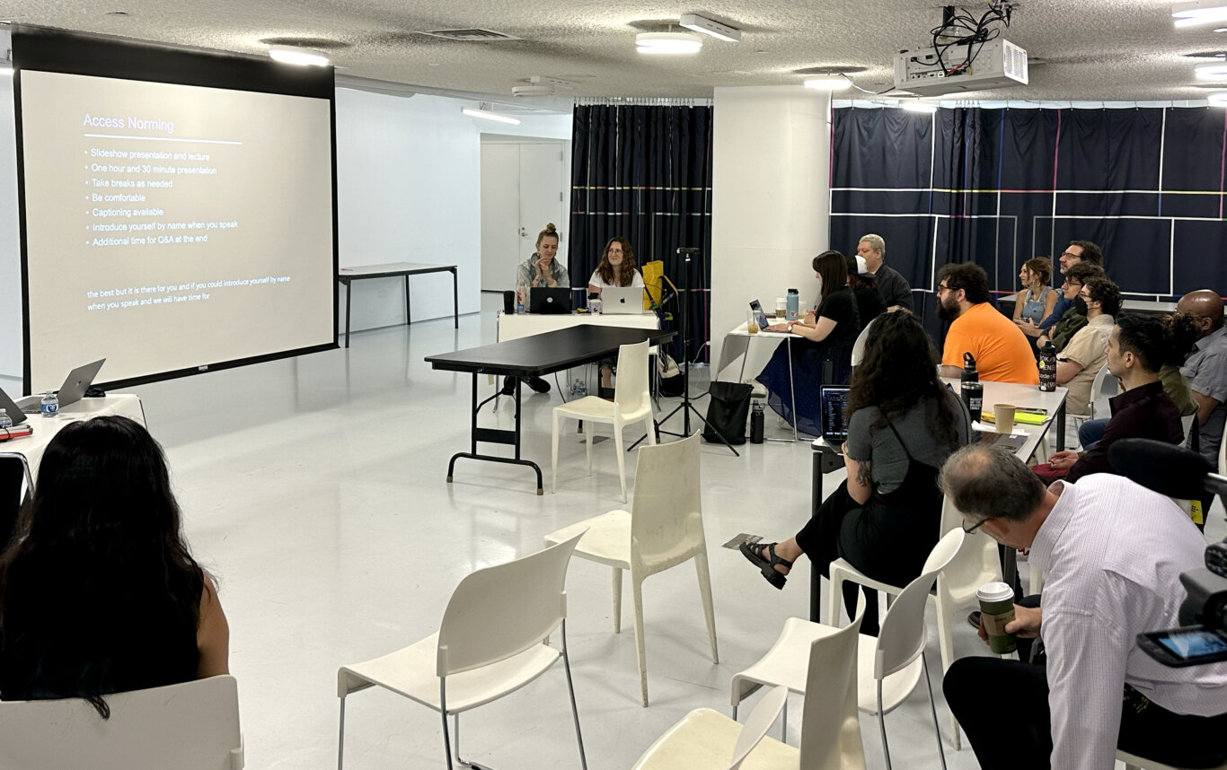 An indoor lecture or conference space with a textured gray ceiling, circular ceiling lights, air vents, and a ceiling-mounted projector. Rows of white chairs and desks are arranged across a glossy white floor. Several attendees are seated, some using laptops or looking toward the front of the room, while a camera operator stands near the right side with filming equipment. Dark curtains with thin colored line patterns cover the walls, and a laptop screen in the foreground appears to show a presentation slide.