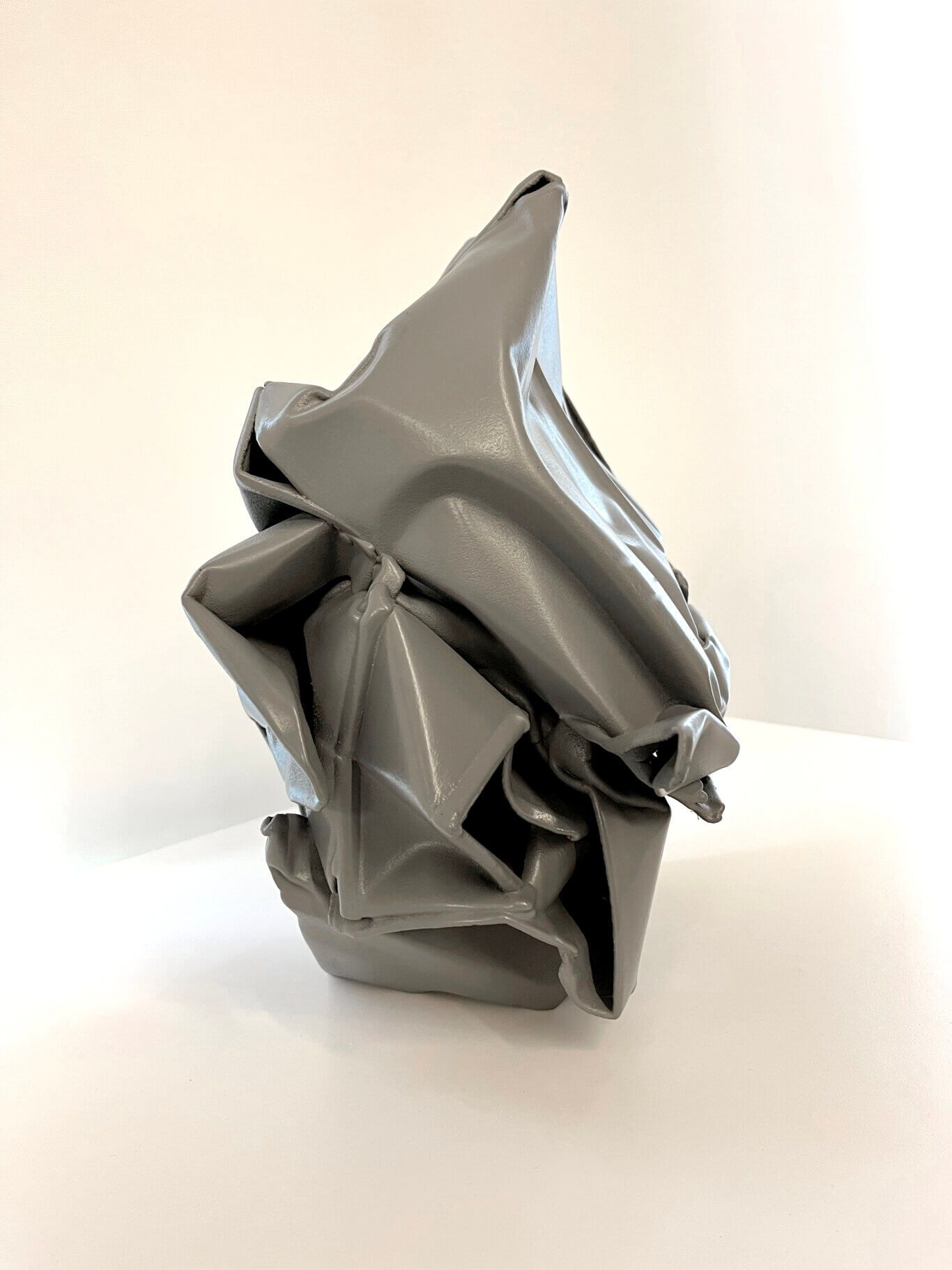 A tightly cropped view of a gray, shiny, crumpled-looking tactile object, resembling folded metal or a crushed bag. It has angular folds, hollow openings, and reflective highlights along its smooth surfaces. The object sits on a white tabletop or shelf, with a softly lit beige wall in the background.