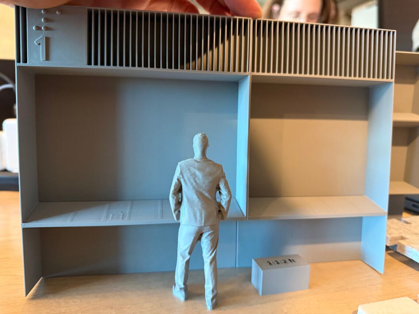 A gray 3D-printed miniature model of a person standing, facing an exhibit display case, with their back towards us. Across the upper row of the case are long rows of thin vertical slats. On the left side, a small raised panel includesthe number one in print and Braille. Below, there are two large open box-like recessed display areas with roll under access below. A small, simplified adult human figure stands in the foreground, facing away from us, their head reaches about two thirds of the way up the main display areas. On the ground, a small grey box is labeled 1:1:2 ft.
