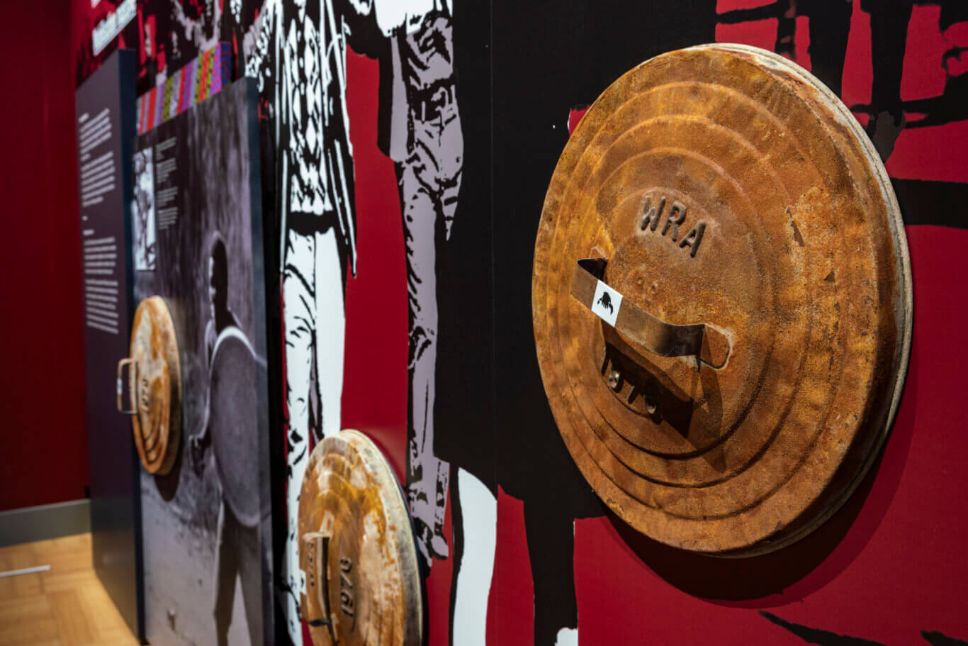 An indoor exhibition display with large rusted, round metal trash can lids attached to a wall. The background panels are deep red and black, decorated with bold black-and-white stencil-like figures and shapes. Some informational text panels are visible but out of focus, suggesting a museum or historical exhibit. The shallow depth of field emphasizes the corroded texture and worn surfaces of the trash can lids while the rest of the display recedes into the background.