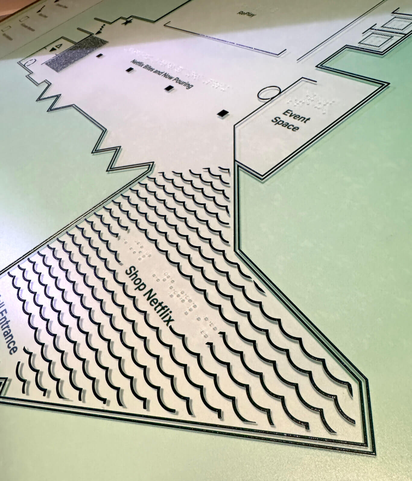 An angled close-up of a glossy, tactile venue map or floor plan. Raised black lines outline different rooms and areas, with printed and Braille labels including "Main Blue and Now Pouring," "Shop Netflix," "Event Space," and "Patio." A large section is filled with repeating curved, scallop-like symbols. The map surface is mostly pale green and white, with strong shadows emphasizing the raised details.