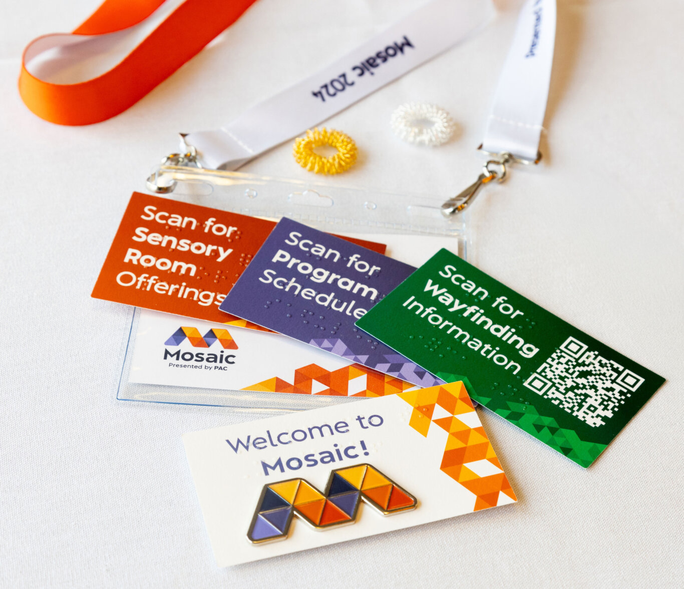A neatly arranged set of Mosaic convening materials on a white fabric surface. Several colorful cards read "Scan for Sensory Room Offerings," "Scan for Program Schedule," and "Scan for Wayfinding Information," with Brailled and QR code. There is a clear plastic badge holder with a red-orange lanyard, a name badge labeled "Mosaic 2024," and a "Welcome to Mosaic!" card featuring a colorful geometric Mosaic logo and enamel-style pin. Small coiled rings in yellow and white, are placed near the badges.