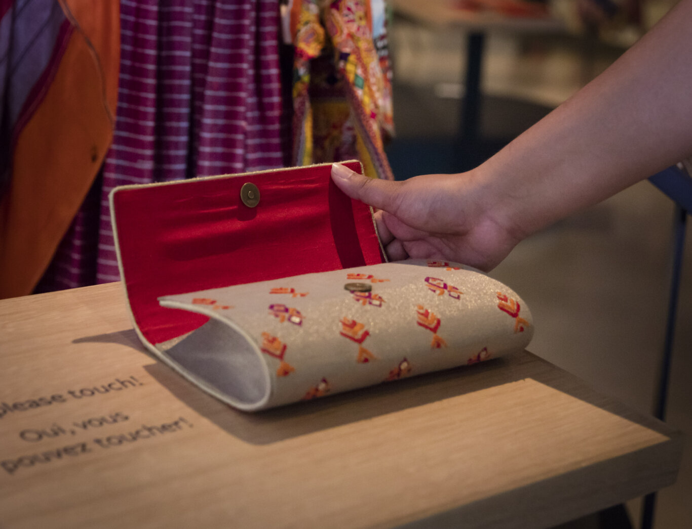 A close-up of a person with medium brown skin tone holding open a silver fabric pouch or purse with red, orange, and purple embroidered geometric shapes on it's surface. The pouch is sitting on a wooden table with the words, "Please touch!" in French and English to the right of the pouch. The inside of the open flap is bright red with a magnetic snap closure. In the background, colorful garments hang or drape, including purple striped fabric and orange material.