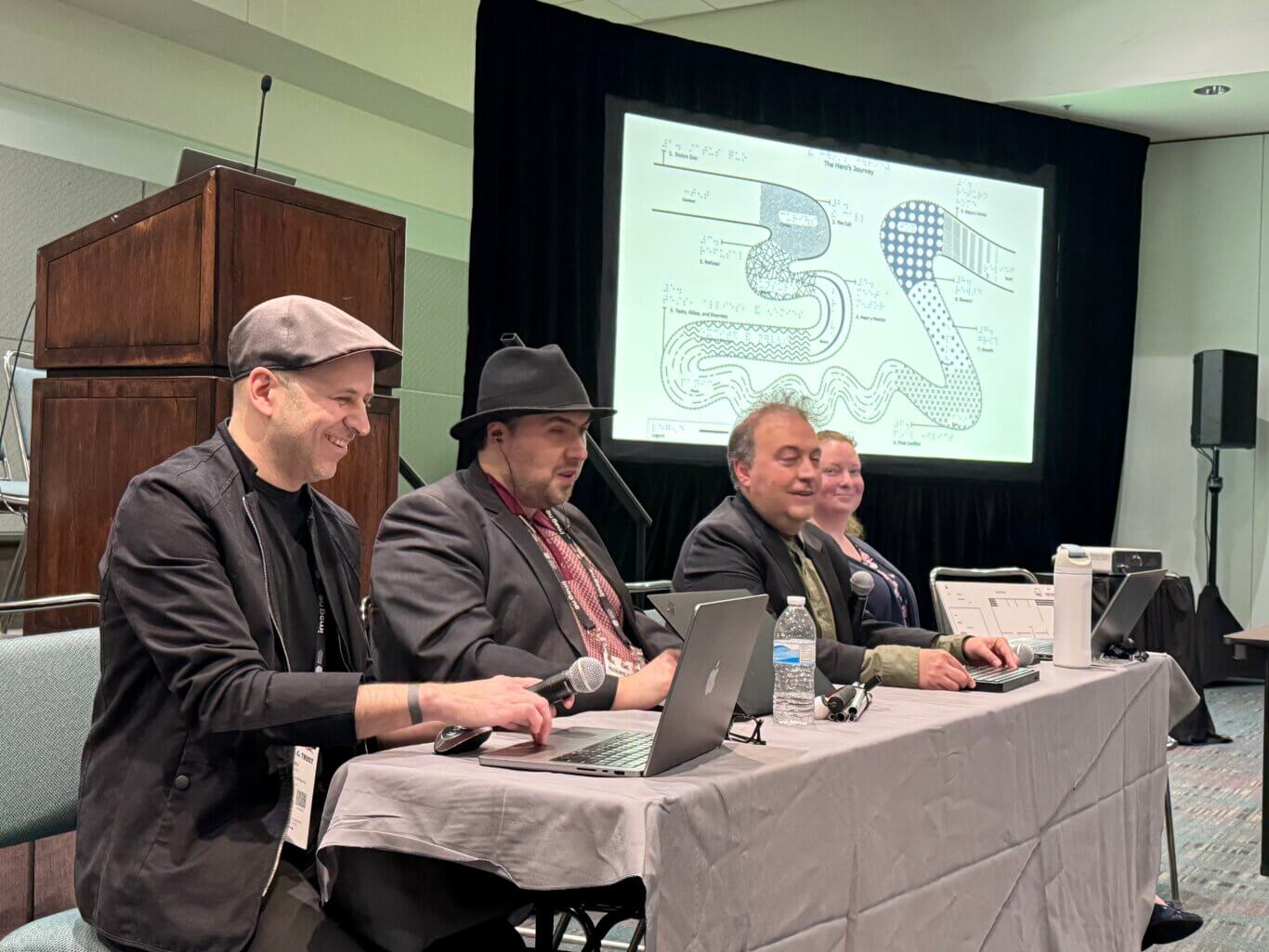 A presentation in a conference room with four smiling people seated at a long table, with microphones, a folded long white cane, laptops, water bottles, and papers in front of them. From our left to right are Corey Timpson, a white man in a grey cap and black jacket, Sina Bahram, a Persian man in a black fedora and black suit jacket, Mark Riccobono, a white man in a black suit coat, and Alison Tyler, a white woman in a navy blue sweater. Corey holds a microphone laughing and typing on a computer. Behind them and to our right, a large projection screen displays a diagram titled "The Hero's Journey."