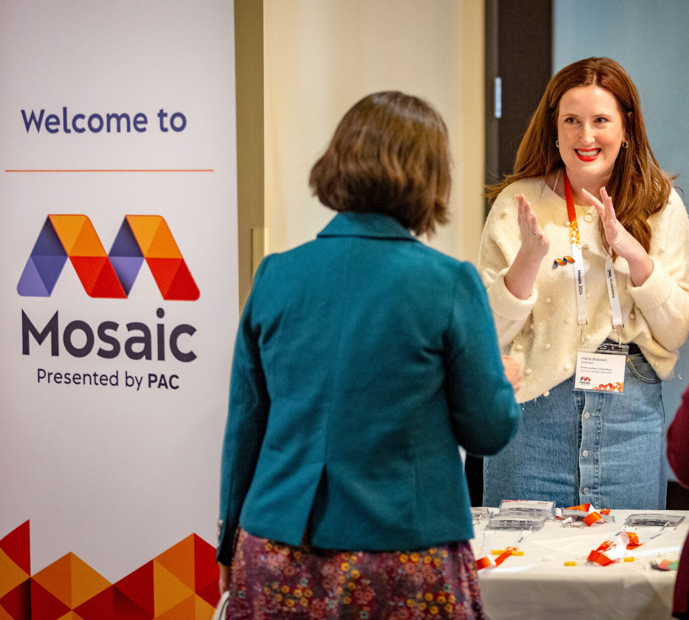 The registration table for Mosaic Convening. Maria Braswell, a white woman on our left, greets Melanie Fales, a white woman, with a large smile and her hands gesturing. On the left is a large vertical sign with the Mosaic logo and words, "Welcome to Mosaic, Presented by PAC." The Mosaic logo is a wide large purple and orange capital M on a white background constructed from 13 equilateral triangles that sits atop three lines of text that say, "Mosaic, Convening 2024, Presented by PAC". "Mosaic" and "Presented by PAC" are rendered in black. A table in front of Maria, holds name badges, lanyards, and event materials.
