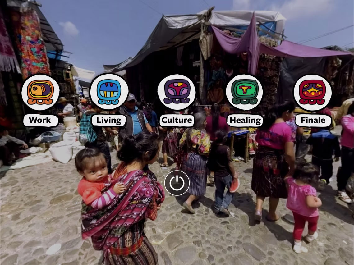 A busy cobblestone market or village street lined with fabric covered stalls, filled with pedestrians and vendors with light brown skin tone in Peru. Many people appear to be wearing colorful patterend traditional woven clothing, including a woman in the foreground carrying a child on her back. Across the middle of the image are five large illustrated circular icons with labels beneath them: "Work," "Living," "Culture," "Healing," and "Finale." A power-button symbol is centered near the bottom, suggesting this is an interactive menu or virtual experience interface layered over a real-world market scene.