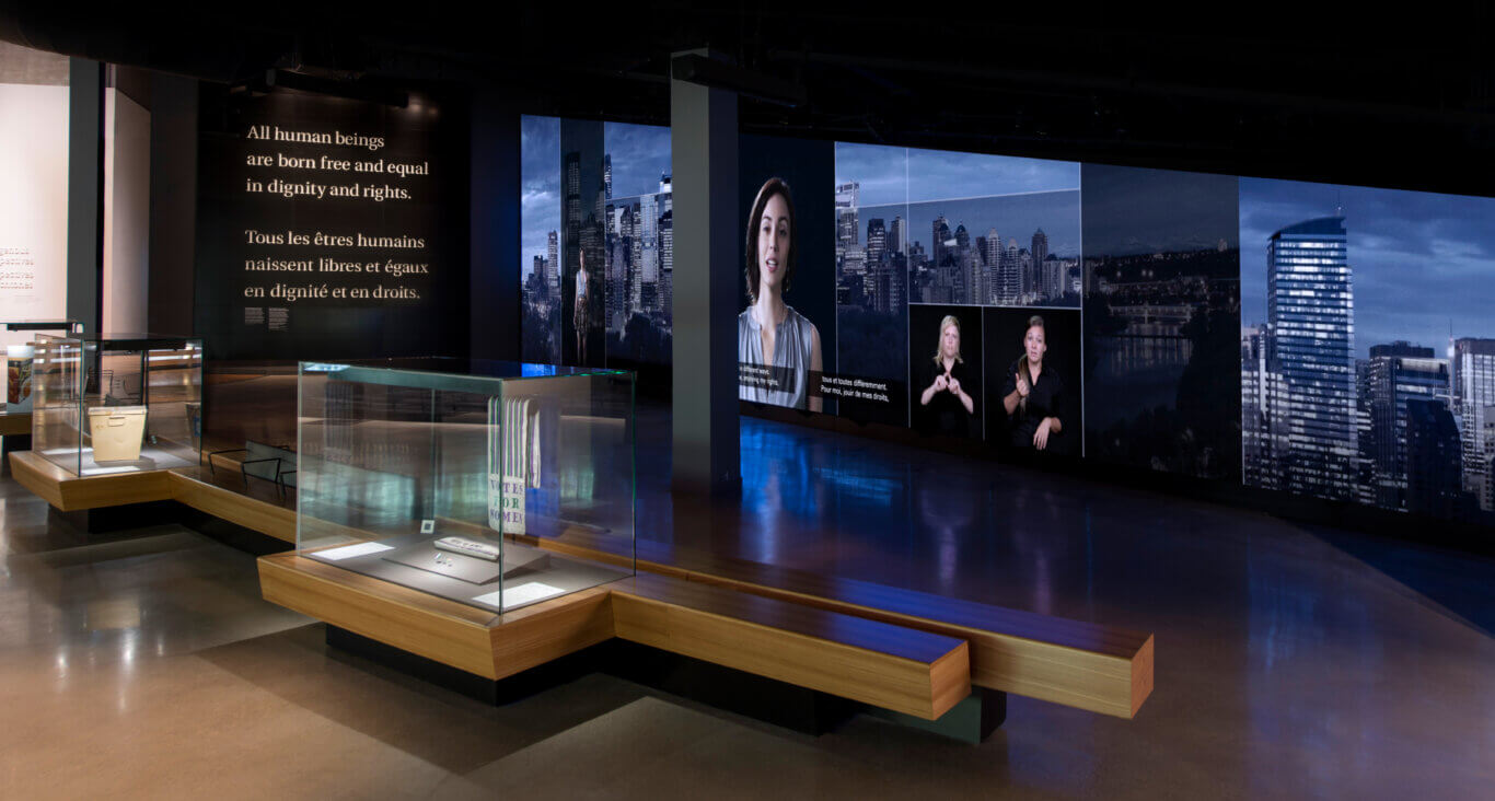 A modern, dark exhibition space with immersive multimedia displays. Large wall screens feature a city skyline, portraits of people, and text from the Universal Declaration of Human Rights, including "All human beings are born free and equal in dignity and rights" in English and French. A video of a person has captions in both French in English with two smaller inset sign language interpreters placed next to the main video. In the foreground are glass display cases set into angular wooden platforms and benches, with reflective polished floors and blue-toned lighting.