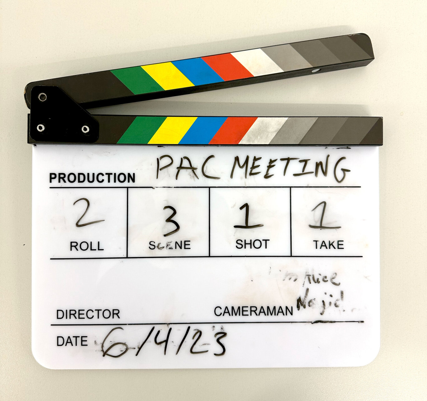 A close-up of a white movie production clapperboard/slate on a light surface. On the slate, the printed label "PRODUCTION" appears on the left, with handwritten text reading "PAC MEETING" across the top field. Below, the slate fields show handwritten numbers: Roll 2, Scene 3, Shot 1, and Take 1. Farther down are printed labels including "DIRECTOR," "CAMERAMAN," and "DATE," with the date handwritten as "6/4/23." The hinged top clapstick is black with diagonal color bands in green, yellow, blue, red, white, and gray. Some marker smudges and partial writing are visible on the right side.