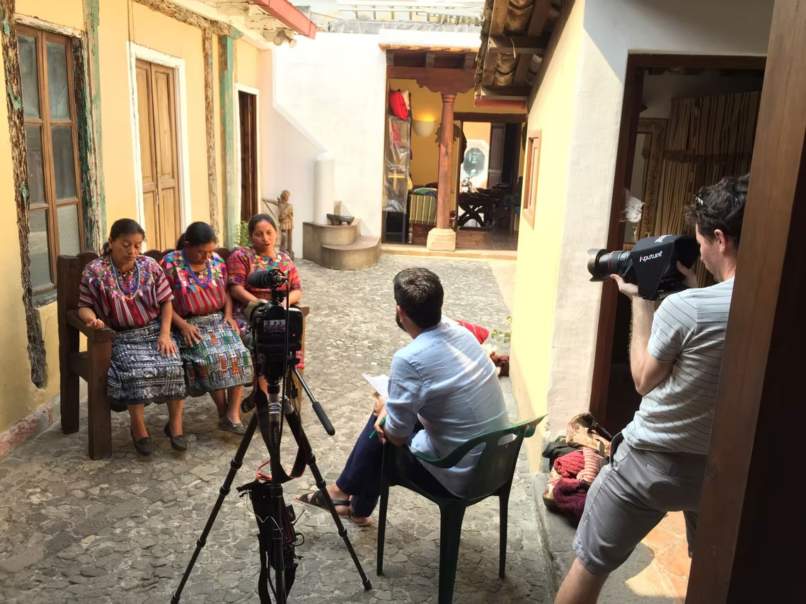 A behind-the-scenes filming setup in a narrow courtyard of three Indigenous Guatemalen women with medium brown skin tone being interviewed by two people with light skin tone wearing business casual clothes. The women are wearing colorful traditional woven blouses and patterned skirts sit together on a wooden bench on our left. A camera on a tripod is positioned in front of them, while a seated interviewer faces them with papers in hand. On the right, another crew member holds a large camera aimed toward the interview scene. The courtyard is cobblestoned, with yellow walls, wooden doors, exposed wooden roof beams, and bright sunlight illuminating the scene.