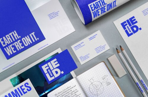 A neatly arranged set of Field Museum brand materials on a gray surface. The design uses a strong cobalt-blue and white color palette with bold, all caps, condensed typography. Visible items include a large folder or brochure reading "EARTH. WE'RE ON IT.", a letterhead sheet, a rolled poster, business cards, an envelope or booklet with the Field Museum logo, two white pencils printed with the campaign slogan, and an open grid notebook containing handwritten notes and a small earth-like sketch.