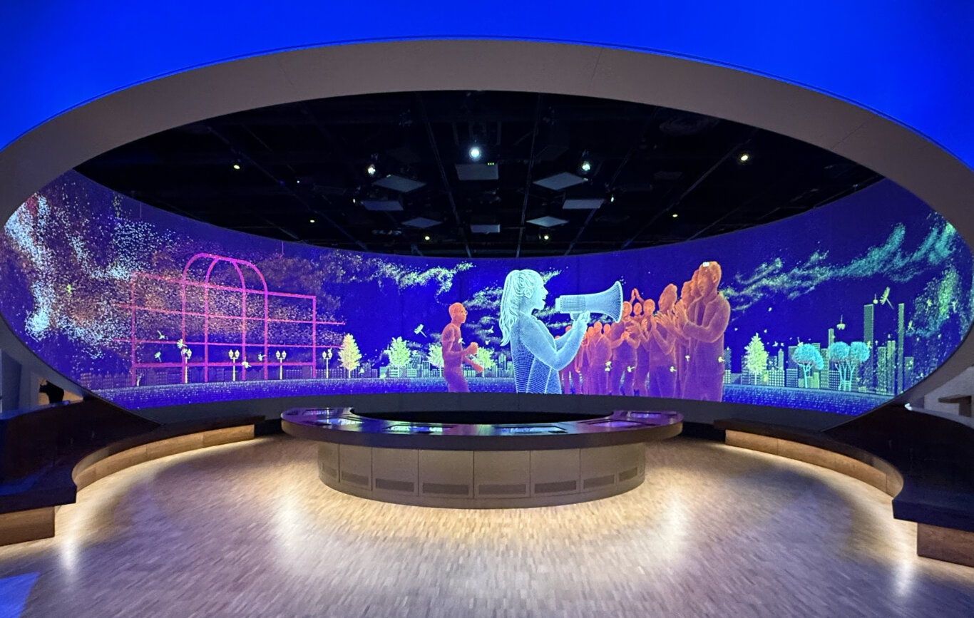 An immersive 270 degree curved theatre digital display filled with vivid neon graphics in blue, purple, green, pink, and orange. The screen appears to show abstract city or park imagery with glowing human-like figures and colorful digital patterns of a person speaking through a bull horn, and a crowd clapping. Above it, sweeping layered ceiling panels glow with intense blue light, while black stage rigging and spotlights are visible overhead. In the middle of the room is a round table with digital interactive screens embedded in the tops. In the foreground, curved seating or platform structures with warm underlighting sit on a pale wood floor.
