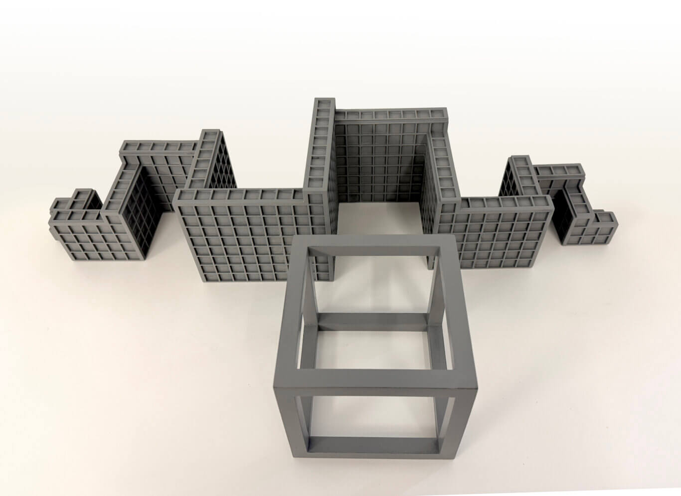 Two geometric tactile objects made of gray rectangular structures. In the foreground, there is a large open frame of a cube. Behind it is a clustered grid of blocks grouped together in a repeating grid of repeated cubes. The scene is photographed from a slightly elevated angle, with a lot of empty white background above the object.