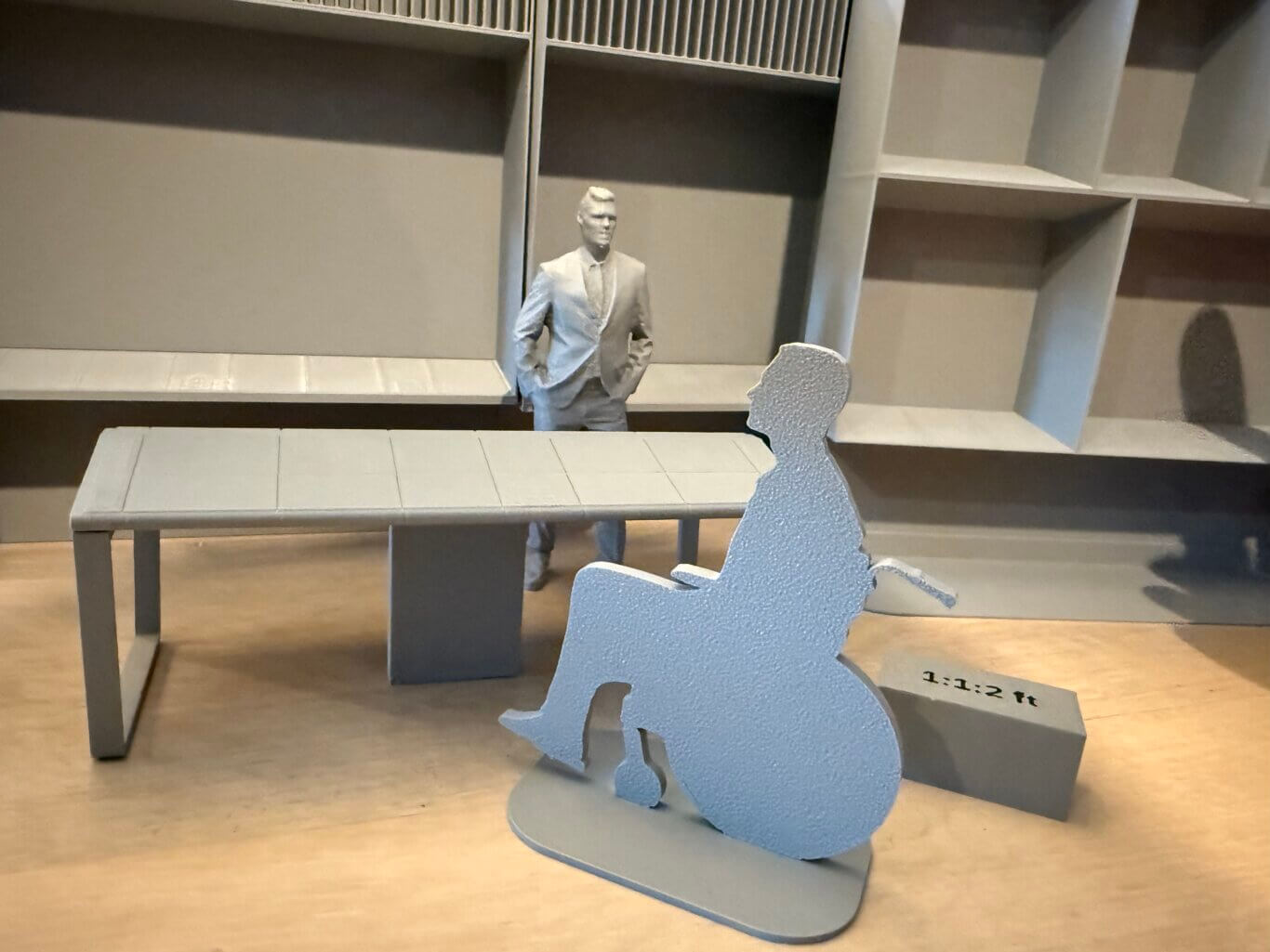 A detailed gray 3D-printed miniature model of a person standing and a person sitting in a wheelchair at a long table, in front of display cases. The small standing figure in a suit is positioned behind the table facing us, while the flat cut out of a person in a wheel chair is pulled up in front of the table. In the background are additional 3D-printed models of multi section display cases. On the ground, a small grey box is labeled 1:1:2 ft.