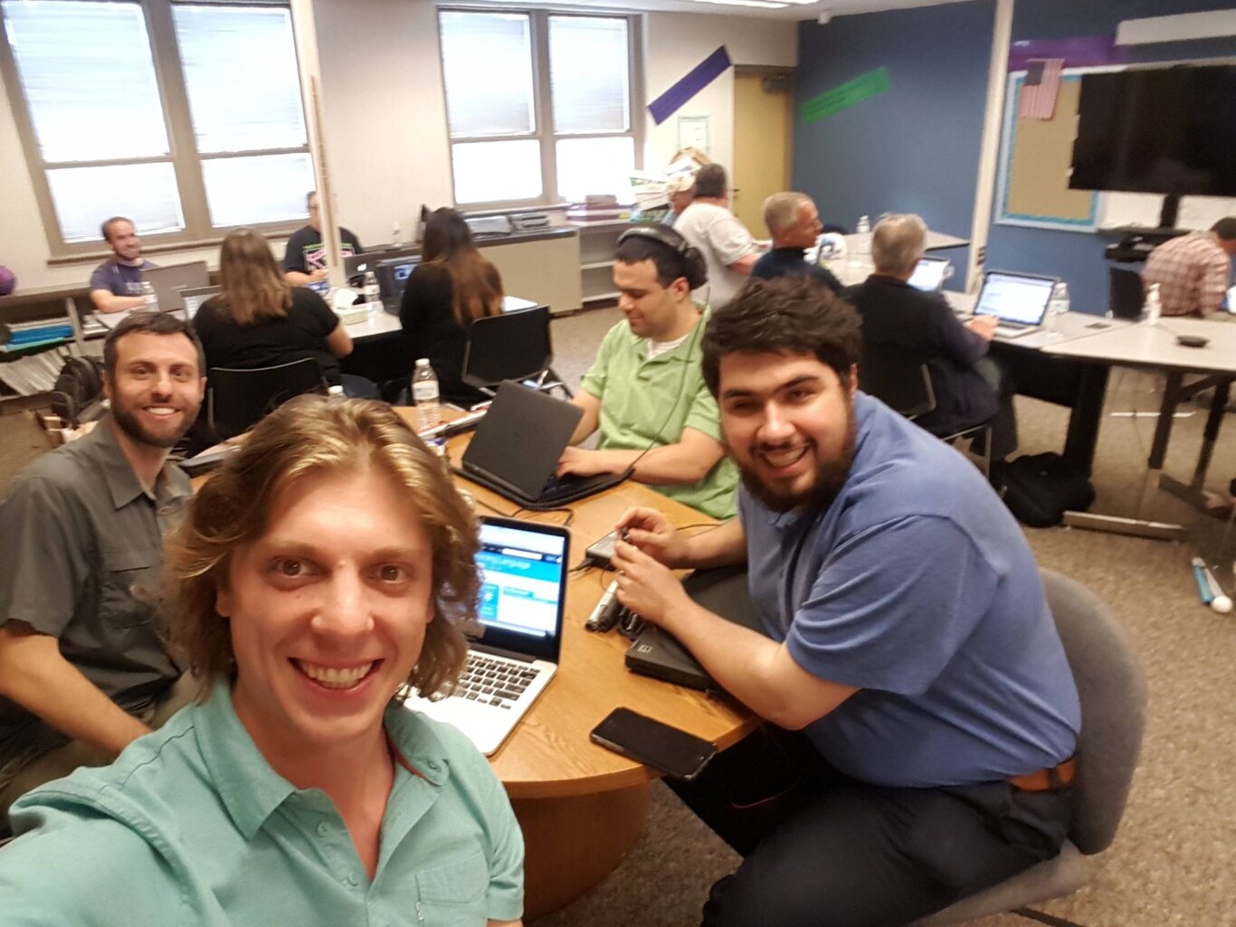 A casual group selfie taken in a classroom of several adults with light skin tone seated around tables using laptops, notebooks, and water bottles. Three people in the foreground are smiling at the camera, including Sina Bahram a Persian man, and Sam Shaw, a white man, while others in the background are working or talking. The room has large windows, wall decorations, a bulletin board, and a large display screen on the right.