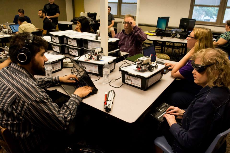 A busy classroom or workshop setting with several people seated around large tables. In the foreground, Sina Bahram, a Persian man wearing headphones uses a laptop while seated near stacked plastic bins and small wheeled robot kits. Across from him, a white woman wearing dark glasses uses a refreshable Braille display. Other participants sit nearby, including a woman at a laptop and a man adjusting his glasses. The tables are covered with electronics, wires, water bottles, robotics components, and storage boxes.