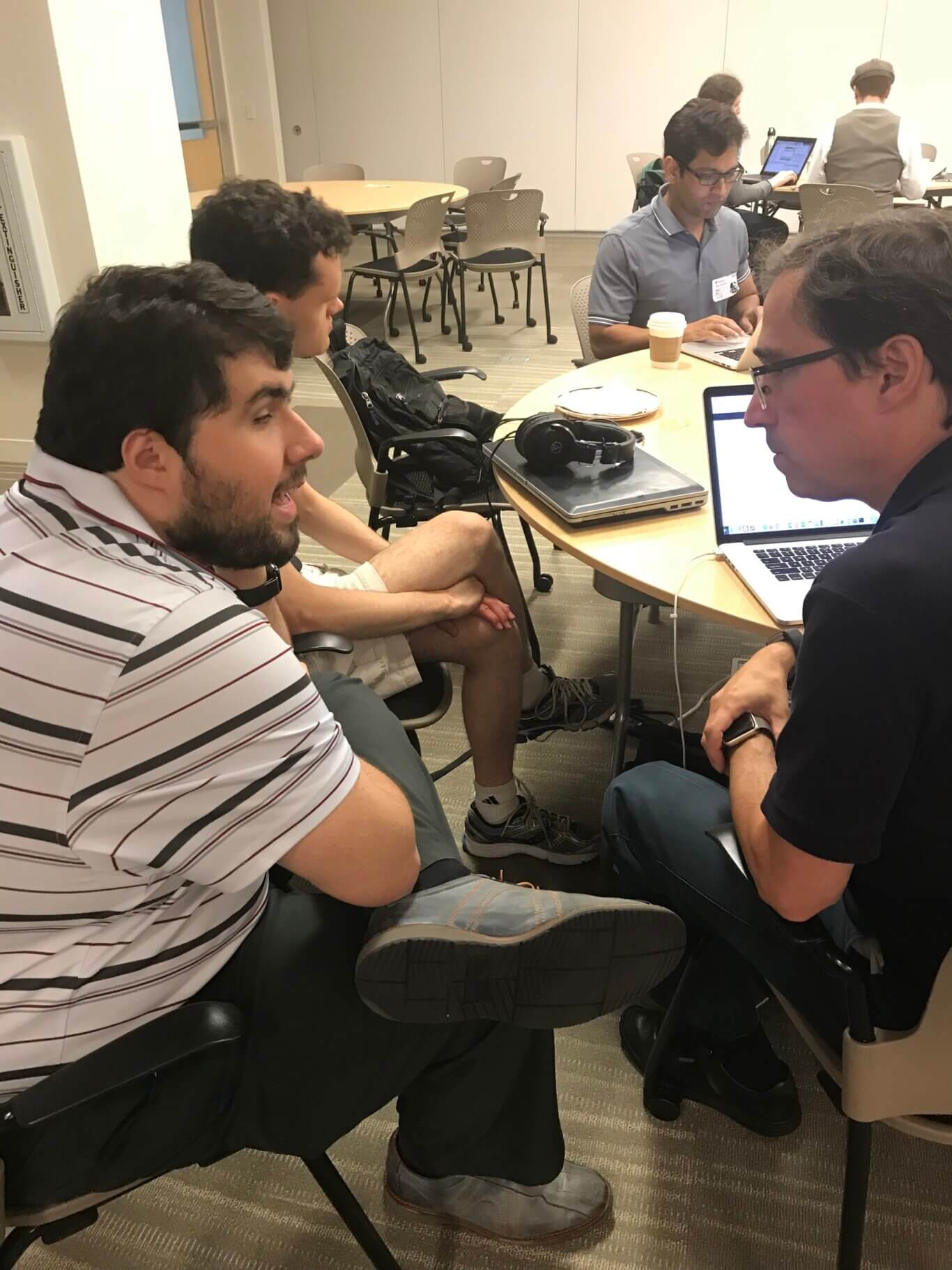 An indoor meeting or study room with several men with medium light skin tone seated around tables. In the foreground, Sina is engaged in conversation. On the table there is a laptop, a pair of over-ear headphones, a coffee cup, papers, and cables. In the background, more people sit at another table using laptops. The room has beige walls, rolling chairs, round tables, carpeted flooring, and a door on the left.