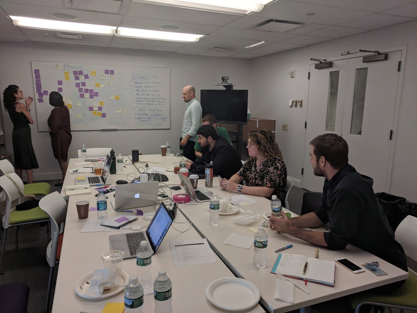 A conference room with seven people collaborating on laptops and at a white board. Several people, including Sina Bahram, a Persian man with light skin tone, are seated around large white tables with laptops, notebooks, water bottles, coffee cups, cables, and papers spread out. At the front of the room, two people stand near a large whiteboard covered with purple, yellow, and white sticky notes, suggesting an ideation or planning activity. The whiteboard includes headings such as "PRODUCT" and "VISITORS," with bullet points written underneath. A wall-mounted monitor and video camera are visible on the right side, and overhead fluorescent lights illuminate the room.