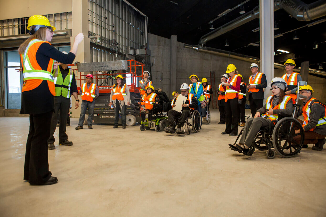 A group of people in a large indoor construction or renovation site with exposed walls, ducts, scaffolding, and unfinished concrete floors. On our left, a person in a yellow hard hat and orange safety vest stands, addressing the group. The attendees, all wearing high-visibility vests and hard hats, are gathered across the space and listening, taking notes, or looking around. Several people are using wheelchairs or other mobility aids.