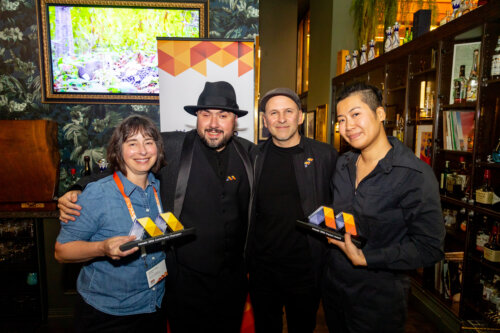 Four people standing close together and smiling at us in a cocktail bar holding trophies in the shape of colorful geometric "M" designs. On our left is Melanie Fales, a white woman with light skin tone who hold the award, "Mosaic 2024 Community Champion." In the middle are Sina Bahram, a Persian man, and Corey Timpson, a white man, both with light skin tone. On our left is Chris Xu, and Asian American, non binary person with light brown skin holding an award with the words, "Mosaic 2024 Impact Award." A vertical banner with orange geometric patterns stands behind the group, matching the design of the awards. In the background of the image are shelves with bottles, floral wallpaper, warm lighting, and a framed television screen showing a bright nature scene.