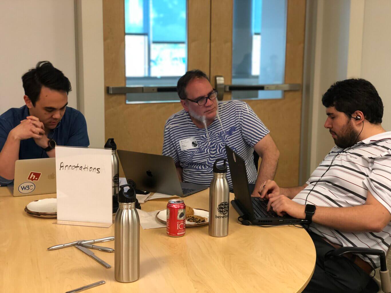 Sina sits around a large wooden table with two men with medium light skin tone. They are focused on laptops and discussion. A white tabletop sign in the foreground reads "Annotations." On the table are multiple laptops, stainless steel water bottles, plates with snacks, utensils, and a red canned drink. In the background are wooden double doors with window panels and blue-tinted light coming through, giving the scene an office or classroom meeting-room atmosphere.