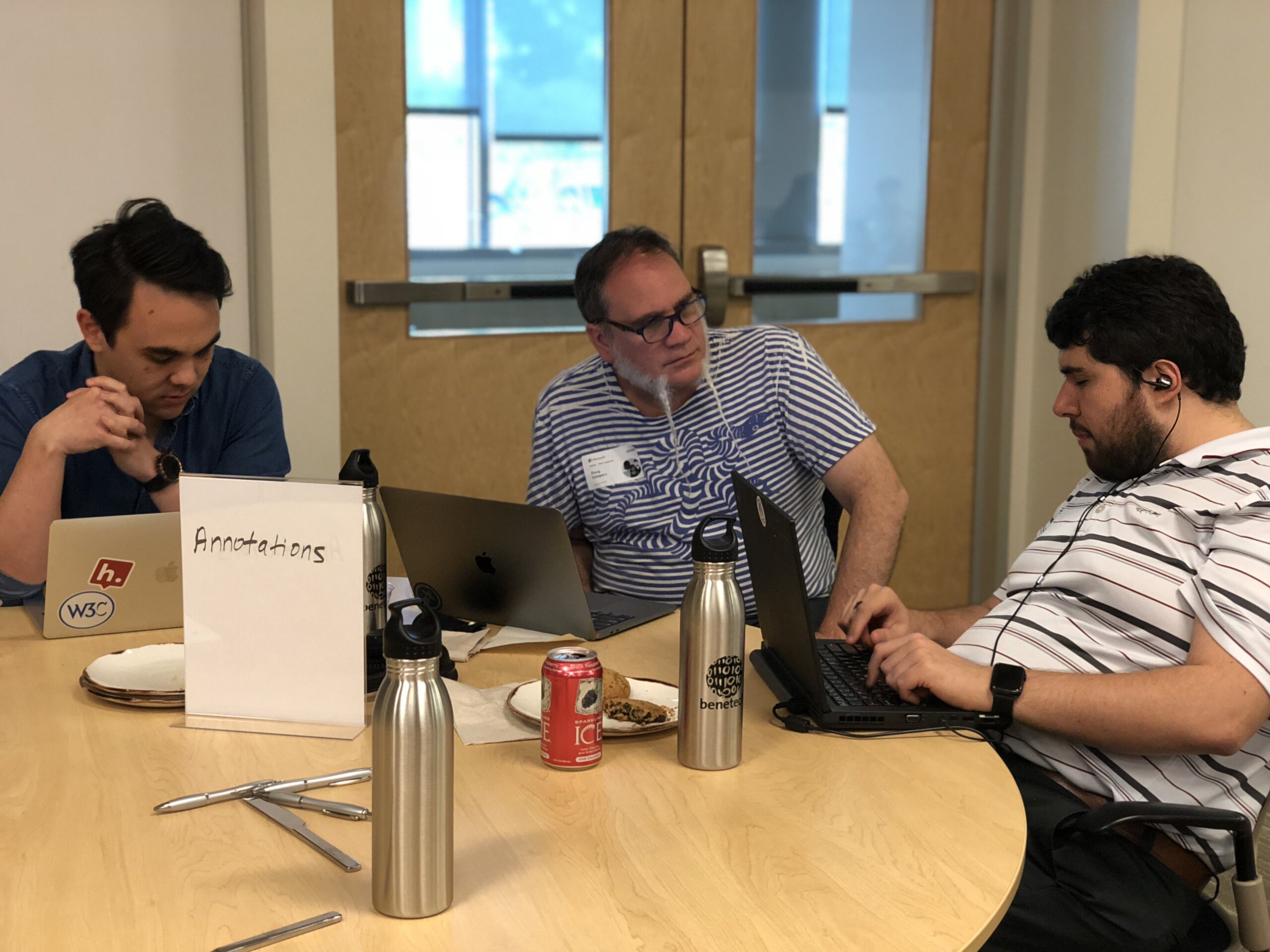 Sina sits around a large wooden table with two men with medium light skin tone. They are focused on laptops and discussion. A white tabletop sign in the foreground reads "Annotations." On the table are multiple laptops, stainless steel water bottles, plates with snacks, utensils, and a red canned drink. In the background are wooden double doors with window panels and blue-tinted light coming through, giving the scene an office or classroom meeting-room atmosphere.