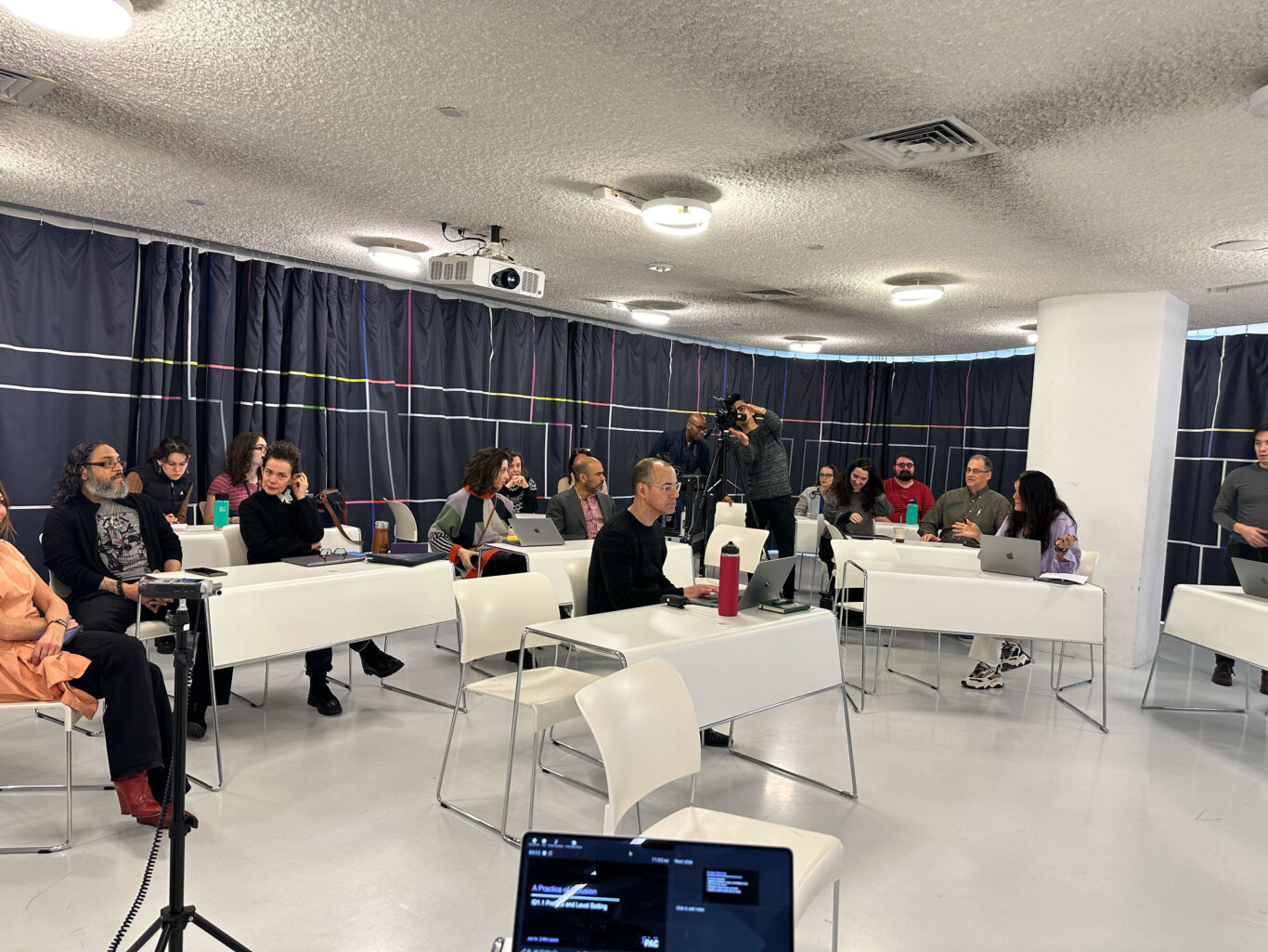 An indoor lecture or conference space with rows of white chairs and desks are arranged across a glossy white floor. The attendees are a mix of genders and races with light to medium dark skin tones, wearing business casual clothing. Several attendees are seated, some using laptops or looking toward the front of the room, while a camera operator stands near the right side with filming equipment. The room has a textured gray ceiling, circular ceiling lights, air vents, and a ceiling-mounted projector. Dark curtains with thin colored line patterns cover the walls, and a laptop screen in the foreground appears to show a presentation slide.