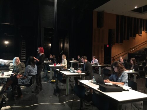The stage of a darkend theatre full of rows of white table tops and 12 people with light to medium skin tone seated at them working on laptops, some alone, and some in groups of two. The room is dimly lit with black stage curtains and equipment on our left, and auditorium seating and wooden wall panels on our right. Cables run across the floor and the tables are full with backpacks, drinks, and keypads.
