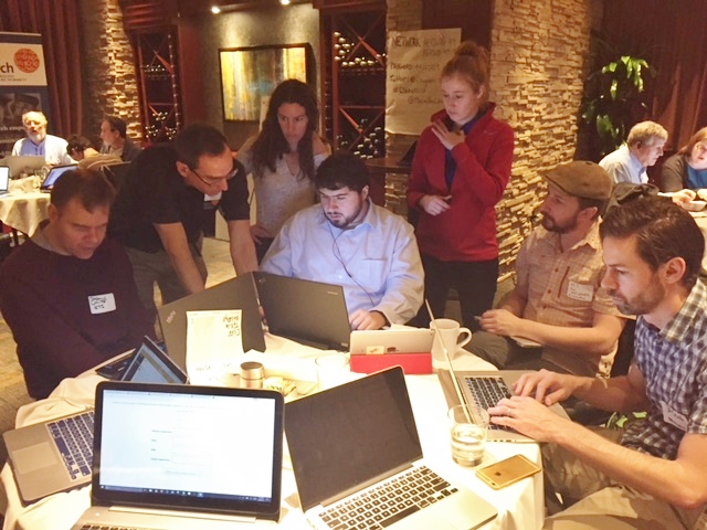 Sina and six adults with light skin tone gathered around a table working collaboratively on laptops. The setting appears to be a cafe or restaurant with warm lighting, stone walls, and wine shelves in the background. They wear business casual clothes and name tags. Multiple laptops, coffee cups, notebooks, and phones are spread across the table. Some people are seated and typing, while others stand or lean in around one laptop.