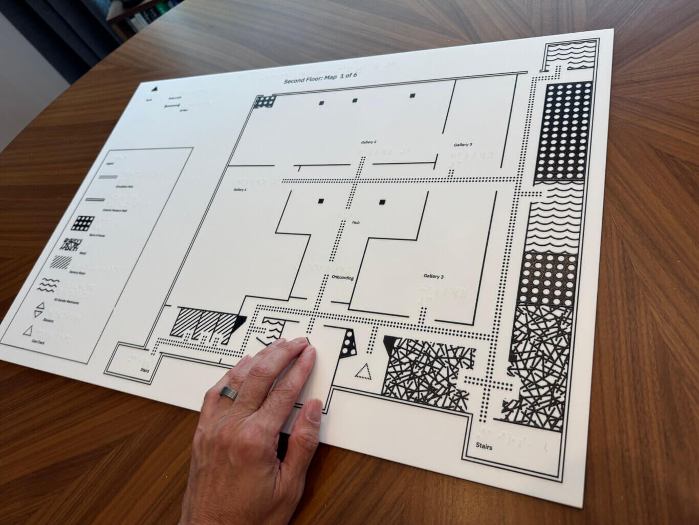 A large tactile floor-plan map printed in black on a white board, placed on a wooden tabletop. The map is labeled "Second Floor: Map 1 of 6" and includes raised braille-like dots, thick outlines, and textured patterns for accessibility. Several areas are labeled, including "Gallery 1," "Gallery 2," "Gallery 3," "Hub," "Onboarding," and "Stairs." A legend on the left side lists symbols for features such as circulation paths, museum walls, entrances, restrooms, elevators, and coat check. At the bottom center, a hand with light skin tone wearing a ring is touching the map.