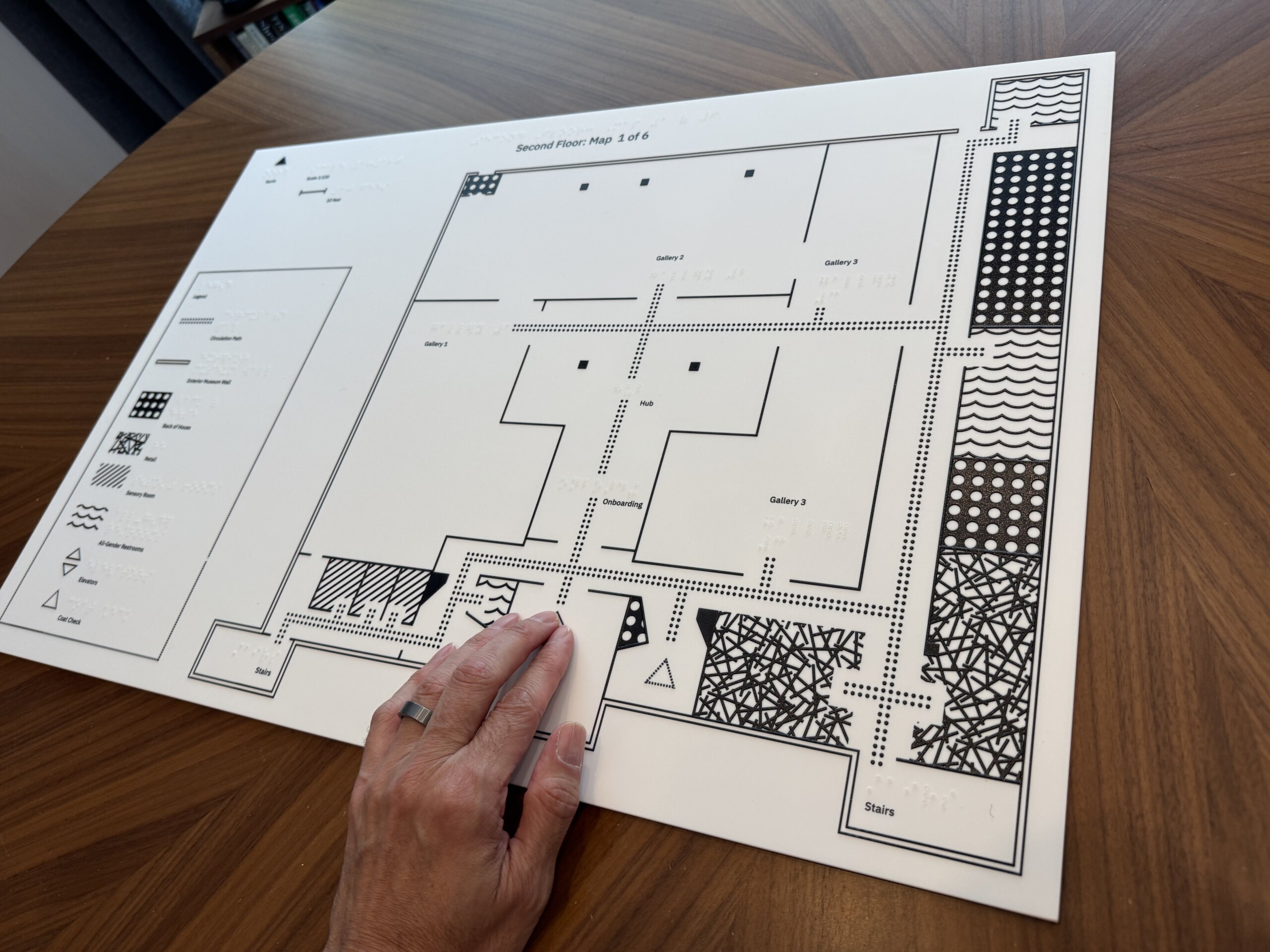 A large tactile floor-plan map printed in black on a white board, placed on a wooden tabletop. The map is labeled "Second Floor: Map 1 of 6" and includes raised braille-like dots, thick outlines, and textured patterns for accessibility. Several areas are labeled, including "Gallery 1," "Gallery 2," "Gallery 3," "Hub," "Onboarding," and "Stairs." A legend on the left side lists symbols for features such as circulation paths, museum walls, entrances, restrooms, elevators, and coat check. At the bottom center, a hand with light skin tone wearing a ring is touching the map.