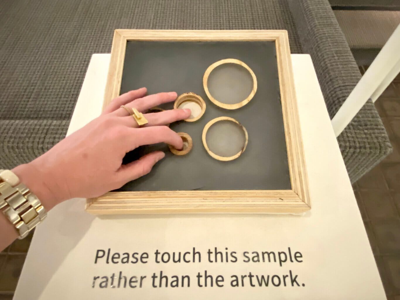 A close-up of a person's hand with light skine tone touching a framed tactile sample board. The board has a dark grey surface with a light wood frame and several round wooden-edged openings or textured circular pieces. It rests on a white surface with partially visible text that reads, "Please touch this sample rather than the artwork." The person wears two gold rings and a thick gold watch band.