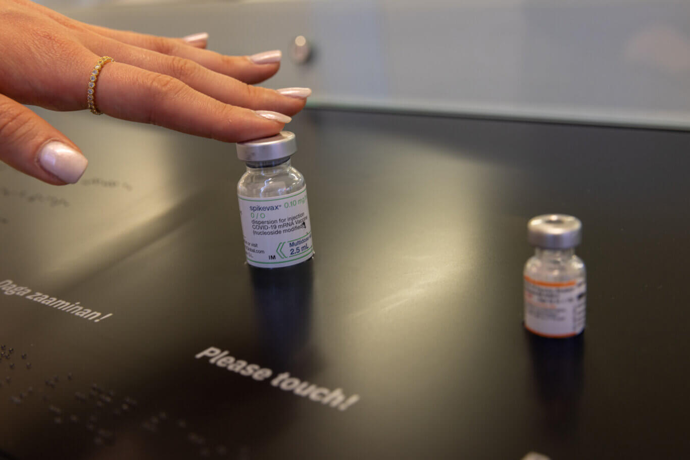 A close-up of a person's hand with medium skin tone touching the metal cap of a small vaccine vial. The hand has light pink polished nails and a delicate gold ring with small stones. The vial label reads "spikevax" and references a COVID-19 mRNA vaccine, with "0.10 mg" and "2.5 mL" visible. Next to it, on our right is a smaller blurred vial. Underneath them, printed text says "Please touch!" and "Daga Zaaminan!" with Braille underneath the print.