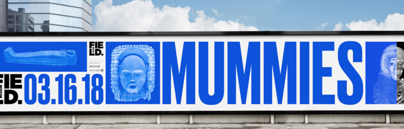 A wide outdoor billboard promoting a museum exhibition titled "MUMMIES." The design uses a stark white background with vivid blue and black typography in all caps. Huge blue letters spell "MUMMIES" across most of the sign, with the date "03.16.18" displayed prominently on the left. Several blue-tinted images of mummified remains and ancient artifacts appear across the billboard, including a wrapped mummy-like figure and a mask or sarcophagus-style face. A small museum logo and "FIELD" branding are visible near the left-center. The billboard is mounted along a street or building facade, with a modern glass building and blue sky with clouds above it.