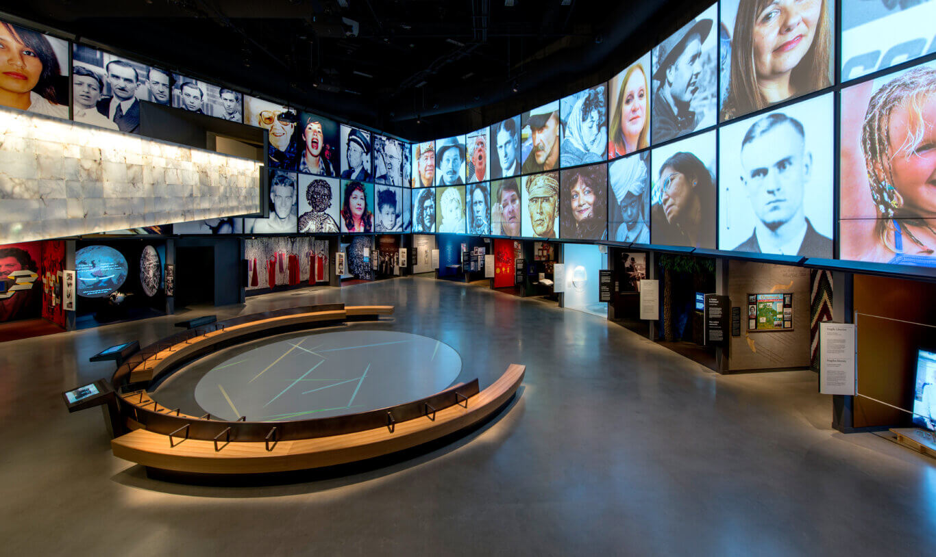 A modern, dark exhibition space with immersive multimedia displays. Large wall screens feature a city skyline, portraits of people, and text from the Universal Declaration of Human Rights, including "All human beings are born free and equal in dignity and rights" in English and French. A video of a person has captions in both French in English with two smaller inset sign language interpreters placed next to the main video. In the foreground are glass display cases set into angular wooden platforms and benches, with reflective polished floors and blue-toned lighting.