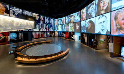 A modern, dark exhibition space with immersive multimedia displays. Large wall screens feature a city skyline, portraits of people, and text from the Universal Declaration of Human Rights, including "All human beings are born free and equal in dignity and rights" in English and French. A video of a person has captions in both French in English with two smaller inset sign language interpreters placed next to the main video. In the foreground are glass display cases set into angular wooden platforms and benches, with reflective polished floors and blue-toned lighting.