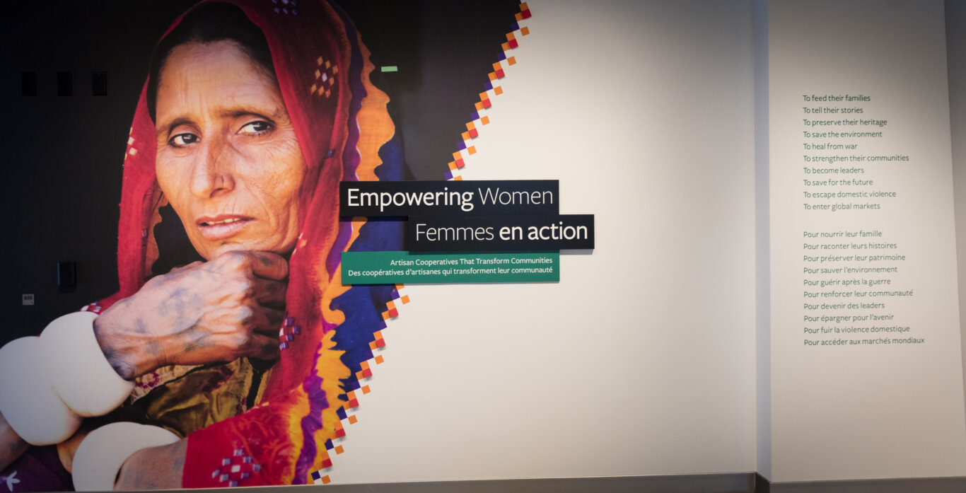 A large exhibition title wall for the show, "Empowering Women," with a floor to cieling colorful photograph of a woman with light golden brown wrinkled skin, partially covered by a red scarf or head covering with bright orange and purple patterned edges. She looks directly at us, her tattooed hand underneath her chin, Large thick white bangles adorn her arms. The sub title "Artisan Cooperatives That Transform Communities," is underneath the main title. All text is in French and English. On our right, a vertical list of green text reads, "To feed their families, to tell their stories, to preserve their heritage, to save the environment, to heal from war, to strengthen their communities, to become leaders, to save for the future, to escape domestic violence, to enter global markets."
