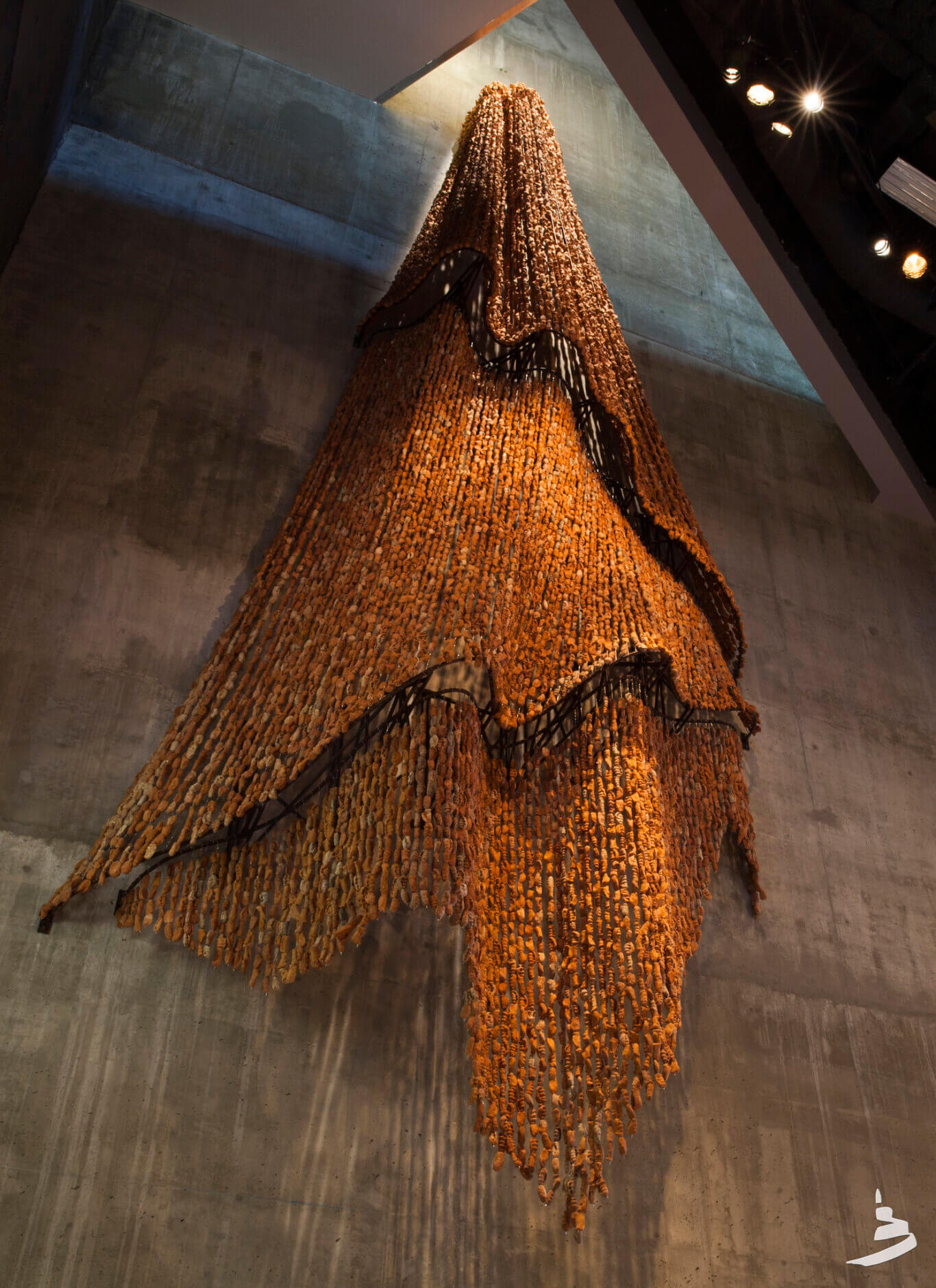 A towering indoor art installation displayed against a high, gray concrete wall of hundreds of clay beads. The piece resembles a cascading textile or curtain, composed of countless vertical strands of hand sized clay beads. It hangs from near the top of the wall and flows downward in layered, irregular waves, with black structural supports or rail-like forms weaving through parts of it. Overhead spotlights illuminate the sculpture, creating warm highlights, deep shadows, and a sense of height and texture within a museum or gallery-like space.