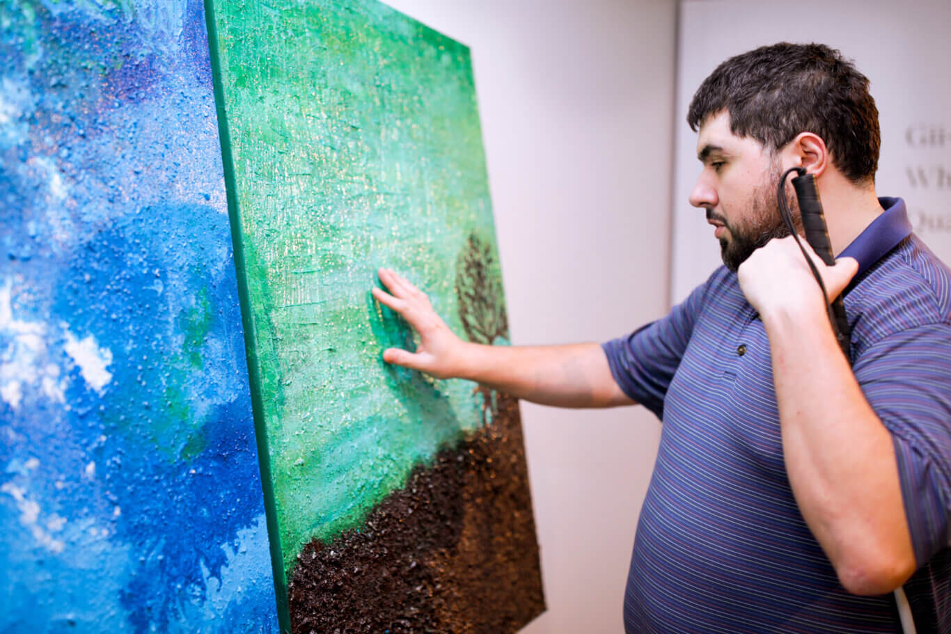 Sina Barham, a bearded Persian man with light skin, in a striped shirt touching several large, glossy, textured canvases. The canvases are painted in vivid colors, with a bright bluepiece on our left and a green and brown piece in front of him. Sina is in profile on our right, with his right hand lighlty touching the canvas surcaface and a long white can tucked into his right elbow.
